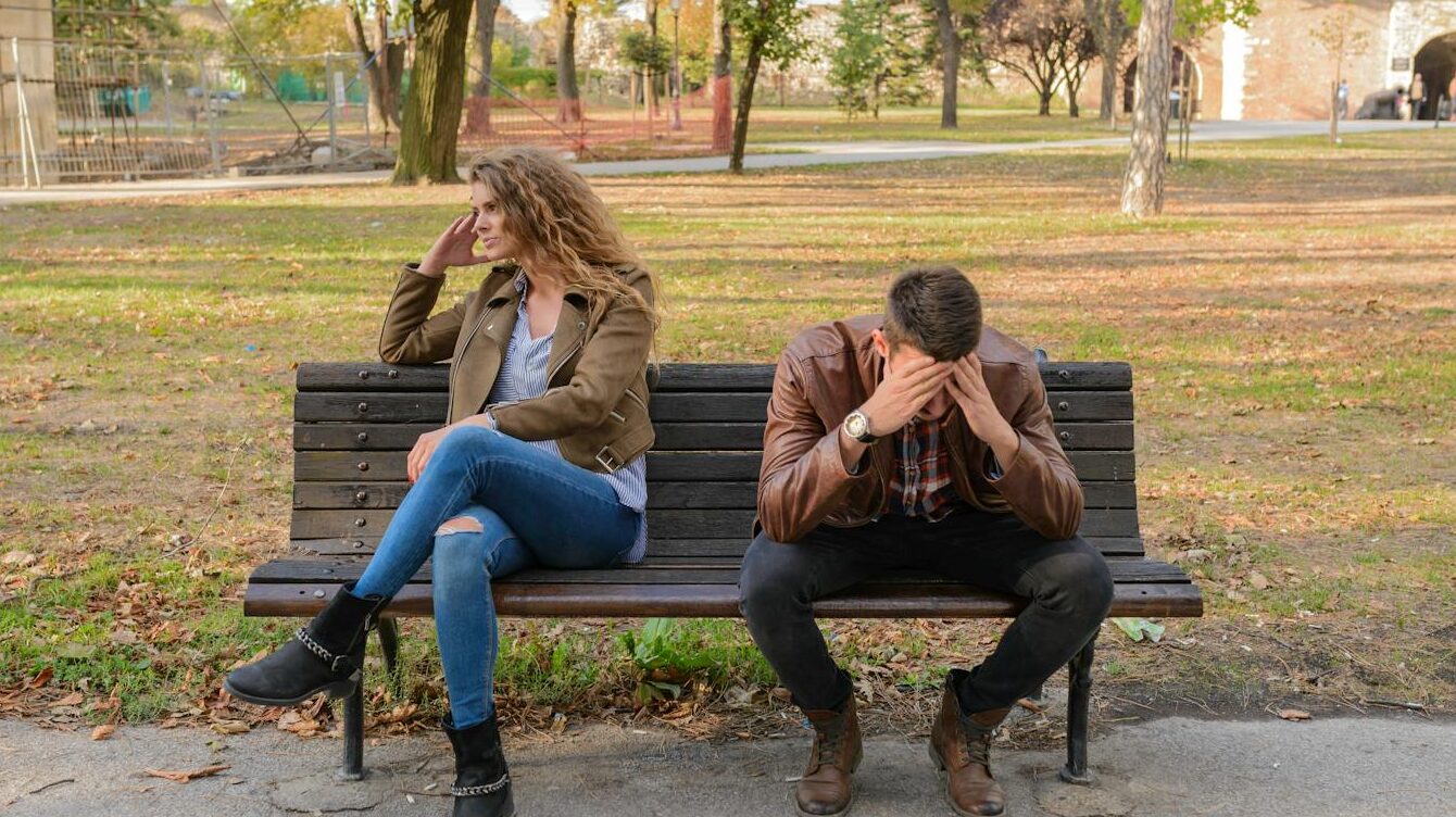 An upset couple seated on a park bench, expressing frustration during an autumn day.